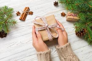 Woman hands give wrapped christmas handmade present in paper with pink ribbon. Present box on holiday on white wooden table, top view photo