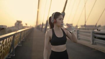 Asian athlete lady exercises drinking water because feel tired after running in urban environment. work out wearing sports clothes on walkway bridge in early morning. video