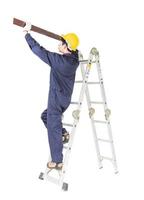 Handyman in uniform standing on ladder holding wood plank on white photo