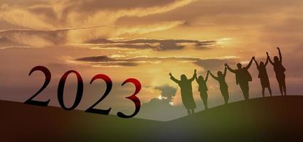 Happy new year 2023, Keep fighting together, Silhouette of 2023 letters on the mountain with business people raised arms in teamwork concept at sunrise. photo