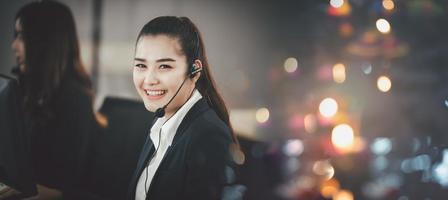 Asian woman in headset, sitting in office, working as operator of call center or support customer service, looking directly at the camera and smiling friendly. photo