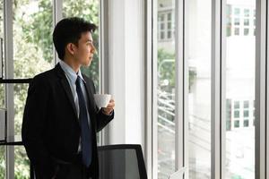 Thoughtful young businessman drinking coffee, looking through window, having break, deep in thoughts, enjoying view, waiting for meeting to start, making business decision. photo