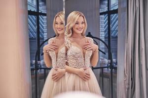 Feeling happy.  Reflection of beautiful young woman wearing wedding dress and smiling while standing in front of the mirror in bridal shop photo