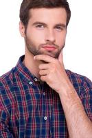 Generating fresh ideas. Portrait of thoughtful young man in casual shirt holding hand on chin and looking at camera while standing isolated on white photo