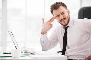 Depressed and frustrated. Depressed young man in formalwear touching his temple with finger gun and keeping eyes closed while sitting at his working place photo