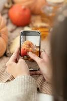 Girl taking photo of pumpkins, autumn leaves and teapot on a window. Girl photographing on phone rustic halloween composition. Happy Thanksgiving and Halloween