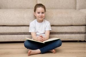 cute little girl sitting at home on the floor barefoot holding a book on her knees and smiling, concept of reading learning back to school photo