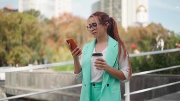 skön företag kvinna i glasögon och en modern turkos jacka använder sig av mobil telefon textning på gata. framgångsrik lady Nästa till företags- kontor skriver text meddelande på smartphone. långsam rörelse. video
