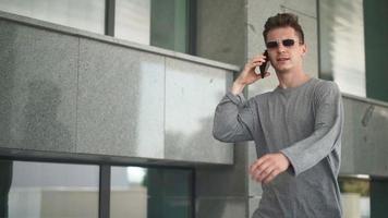 Handsome Caucasian man with in sunglasses walks and speaks on a mobile phone in a city park. Young happy guy laughing and smiling while talking on the phone outdoors. Slow motion. video