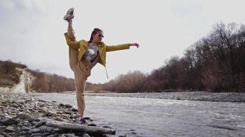 chica atlética con un buen estiramiento se divide contra el telón de fondo de un río de montaña y rocas. la mujer practica yoga y se estira en el fondo de un hermoso paisaje natural. camara lenta. video