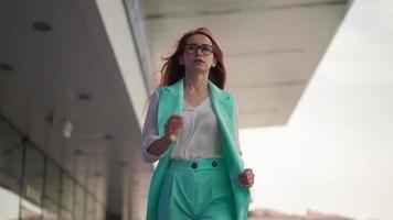 skön självsäker företag kvinna i glasögon och en turkos modern kostym promenader. en ung kvinna entreprenör med lång röd hår promenader genom stadens centrum nära kontor. långsam rörelse. video