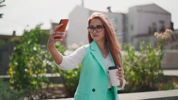A beautiful modern woman in glasses and a fashionable turquoise suit with long red hair and coffee in her hands makes a selfie using mobile phone against the backdrop of city buildings. Slow motion