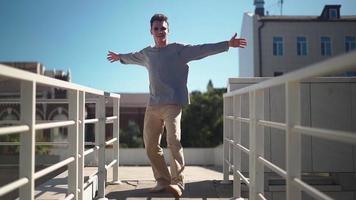 Handsome happy man in sunglasses is actively running, smiling and dancing with joy and happiness along the city street. Scene Shot in an Urban Park Next to Business Center. Slow motion. video