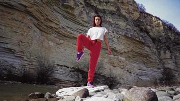 Athletic girl with a good stretch does splits against the backdrop of a mountain river and rocks. Woman practices yoga and stretching on the background of a beautiful natural landscape. Slow motion. video