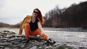 Schöne modische Frau mit Sonnenbrille, die am felsigen Ufer eines Gebirgsflusses ruht. Lebensstil und Reisen. Zeitlupe. video