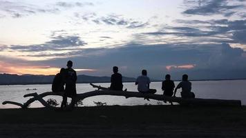 gorontalo, indonésia, outubro de 2022 - silhueta de um grupo de jovens relaxando à beira do lago enquanto aprecia o pôr do sol video