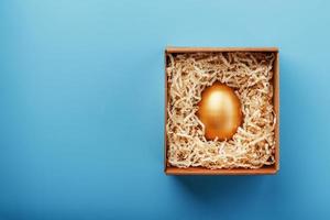 Golden egg in a cardboard box with shavings on a blue background.