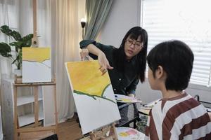 Female Asian teacher teaches and demonstrates to the student boy on acrylic color picture painting on canvas in art classroom, creatively learning with skill at the elementary school studio education. photo