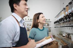 Asian male shopkeeper describes natural organic products to woman customer in refill store, zero-waste and plastic-free grocery, environment-friendly, sustainable lifestyles with reusable containers. photo