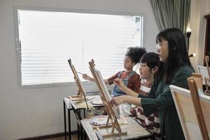 A female Asian teacher teaches and demonstrates to the children on acrylic color picture painting on canvas in art classroom, creatively learning with skill at the elementary school studio education. photo