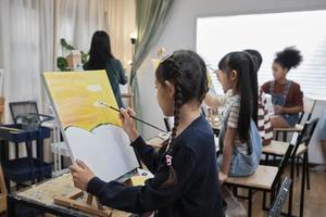 un grupo de niños multirraciales que aprenden con una mujer asiática enseña pintura acrílica en color sobre lienzo en el aula de arte, aprendiendo creativamente con habilidad en la educación de estudio de la escuela primaria. foto