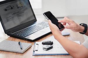 Car insurance online concept , A man hold smartphone and with checklist car key toy car on table and use laptop check online case of accident photo