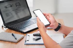 Car insurance online concept , A man hold smartphone and blank white screen with checklist car key toy car on table and use laptop check online case of accident photo