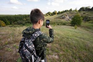Activity on sunny autumn day, boy exploring nature. Kid wear backpack hiking and making video on phone. photo