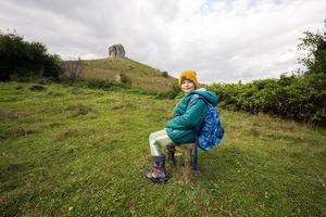 Activity on sunny autumn day, boy exploring nature. Kid wear yellow hat and backpack sit against big stone of Pidkamin, Ukraine. photo