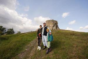 Father with two sons wear backpack hiking near big stone in hill. Pidkamin, Ukraine. photo