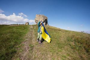 Welcome to Ukraine. Boy hold ukrainian flag near big stone in hill Pidkamin. photo