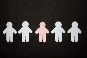 Silhouettes of five men, arranged in a row, cut from paper. Between the white men there is one pink man. On black background photo