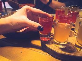 mujer, niña sostiene en la mano con una manicura en los dedos un delicioso vaso rojo, un trago con alcohol fuerte, vodka, brandy, brandy en soportes de madera sobre una mesa en un café, bar, restaurante foto