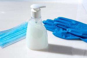 guantes de goma azul, mascarilla médica y botella de jabón sobre un fondo claro. foto