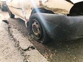 Old white rusty broken car of the carcass with lowered punctured wheels with scratches corrosion and a torn-off bumper with dents and scratches thrown on the road. Auto junk photo