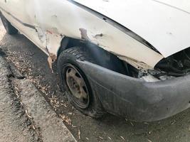 viejo coche blanco oxidado roto de la carcasa con ruedas pinchadas bajadas con arañazos corrosión y un parachoques roto con abolladuras y arañazos tirados en la carretera. chatarra de auto foto