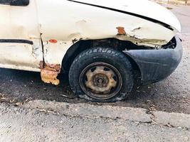 viejo coche blanco oxidado roto de la carcasa con ruedas pinchadas bajadas con arañazos corrosión y un parachoques roto con abolladuras y arañazos tirados en la carretera. chatarra de auto foto