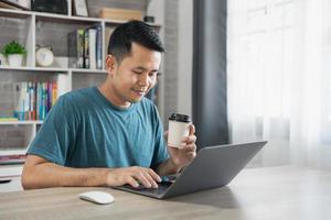 Asian freelance man smiling holding cup of hot coffee and working on laptop computer on wooden table at home. Entrepreneur man working for his business at home. Business work at home concept. photo