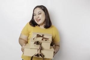 una joven y feliz mujer asiática de gran tamaño lleva una camisa amarilla y sostiene una caja de regalos y una bolsa de compras. foto