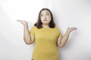 A portrait of an Asian big size woman wearing a yellow shirt shrugging her shoulder looks so confused, isolated by a white background photo