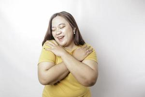 joven y hermosa mujer asiática de gran tamaño con una camiseta amarilla sobre fondo blanco abrazándose a sí misma feliz y positiva, sonriendo confiada. amor propio y autocuidado foto