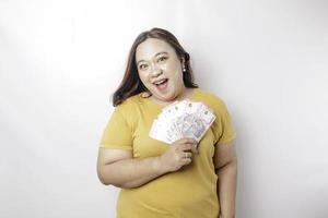 una mujer joven y feliz de gran tamaño lleva una camiseta amarilla y tiene dinero en efectivo en rupias indonesias aislada de fondo blanco foto