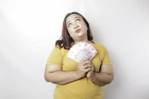 una mujer joven y pensativa de gran tamaño lleva una camiseta amarilla y tiene dinero en efectivo en rupias indonesias aislada de fondo blanco foto
