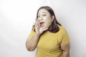 joven y hermosa mujer asiática de gran tamaño con una camisa amarilla gritando y gritando fuerte con una mano en la boca. concepto de comunicación foto