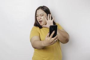 The angry and mad face of big sized Asian woman in yellow shirt while holding her phone on isolated white background. photo