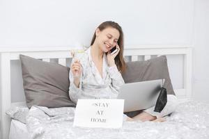 Beautiful happy young woman with a glass of water working on laptop computer in the bedroom. Stay at home. Freelancer. Writing, typing. Girl checking social apps. Communication and technology concept. photo