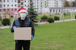 Delivery man in protective mask, gloves and santa's hat holding box in hands outdoors, delivery service during coronavirus in holiday time. photo