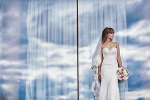 hermosa novia elegante de lujo con un vestido blanco con un ramo en las manos cerca de la ventana panorámica foto