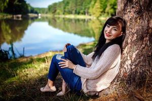 Beautiful young woman in jeans sits on river bank near big tree in sunny day photo