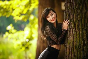 chica morena sexy con blusa de encaje y falda de cuero con tacones altos posando cerca de un árbol en el bosque foto